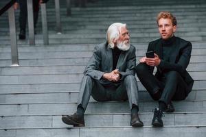 Sitting with phone. Young guy with senior man in elegant clothes is outdoors together. Conception of business photo