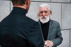 Having a meet. Young guy with senior man in elegant clothes is outdoors together. Conception of business photo