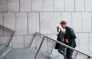 Walking on the stairs. Young guy with senior man in elegant clothes is outdoors together. Conception of business photo