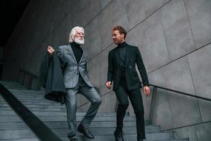 Walking on the stairs. Young guy with senior man in elegant clothes is outdoors together. Conception of business photo