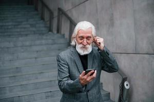 Senior businessman in formal clothes, with grey hair and beard is outdoors trying wireless headphones with phone photo