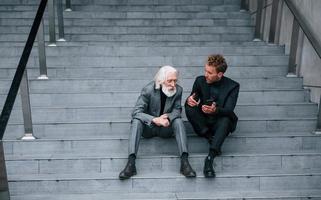 Sitting with phone. Young guy with senior man in elegant clothes is outdoors together. Conception of business photo