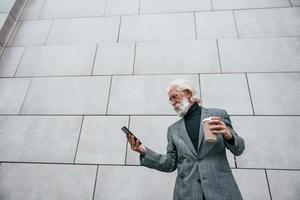 usando un teléfono inteligente. hombre de negocios senior en ropa formal, con pelo gris y barba está al aire libre foto