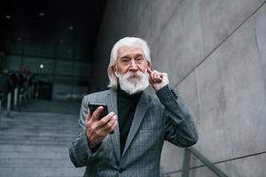 Senior businessman in formal clothes, with grey hair and beard is outdoors trying wireless headphones with phone photo
