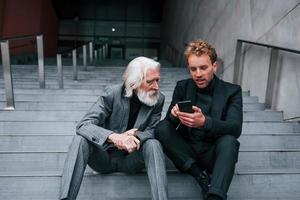 Sitting with phone. Young guy with senior man in elegant clothes is outdoors together. Conception of business photo