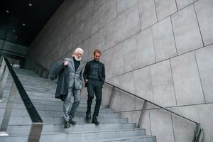 Walking on the stairs. Young guy with senior man in elegant clothes is outdoors together. Conception of business photo