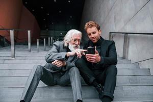 Sitting with phone. Young guy with senior man in elegant clothes is outdoors together. Conception of business photo