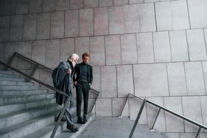 Walking on the stairs. Young guy with senior man in elegant clothes is outdoors together. Conception of business photo