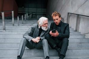 Sitting with phone. Young guy with senior man in elegant clothes is outdoors together. Conception of business photo