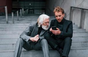Sitting with phone. Young guy with senior man in elegant clothes is outdoors together. Conception of business photo