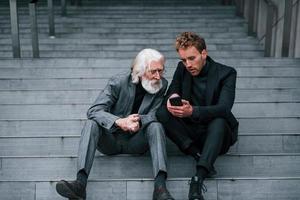 sentado con el teléfono. un joven con un anciano vestido con ropa elegante está juntos al aire libre. concepción del negocio foto