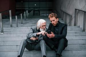 Sitting with phone. Young guy with senior man in elegant clothes is outdoors together. Conception of business photo