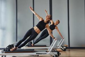 de pie sobre equipos de gimnasia y haciendo estiramientos. dos mujeres con ropa deportiva y con cuerpos delgados tienen un día de yoga en el interior juntas foto