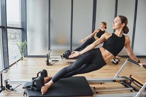 Standing on gym equipment and doing stretches. Two women in sportive wear and with slim bodies have fitness yoga day indoors together photo