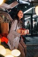 Near the car with gift box. Cheerful woman is outdoors at Christmas holidays time. Conception of new year photo