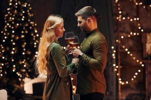 Standing and holding big glasses with wine. Young lovely couple have romantic dinner indoors together photo