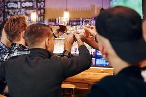 Cheering by knocking beer glasses. Group of people together indoors in the pub have fun at weekend time photo