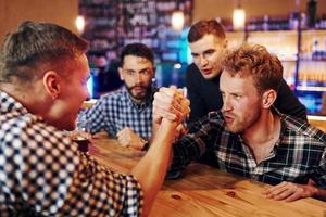 Friends plays arm wrestling. Group of people together indoors in the pub have fun at weekend time photo