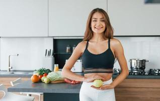 sostiene la manzana contra el vientre. joven europea está en el interior de la cocina con comida saludable foto