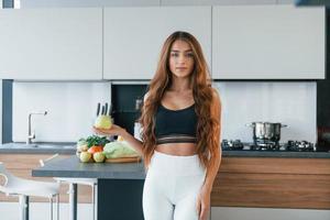 Holds apple in hand. Young european woman is indoors at kitchen indoors with healthy food photo