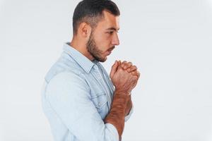 Guy is praying. Young handsome man standing indoors against white background photo