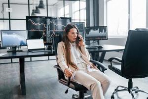 Talks by phone. Young female stock brocker works indoors in the office. Many of displays photo