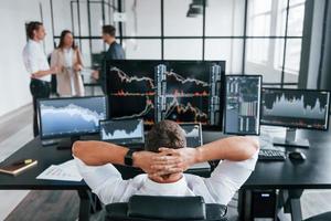 Man sits on chair and takes break. Team of stockbrokers works in modern office with many display screens photo