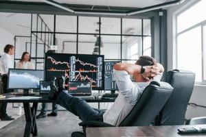 Man sits on chair and takes break. Team of stockbrokers works in modern office with many display screens photo