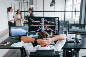 Man sits on chair and takes break. Team of stockbrokers works in modern office with many display screens photo