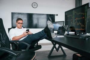 Sits on the chair. Young male stock brocker works indoors in the office. Many of displays photo