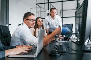 Sitting by the table. Team of stockbrokers works in modern office with many display screens photo