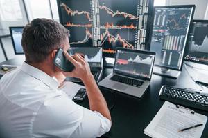 Talks by phone. Young male stock brocker works indoors in the office. Many of displays photo