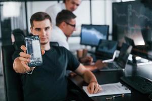 Man shows information that is on phone display. Team of stockbrokers works in modern office with many display screens photo