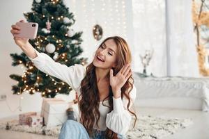 Happy young woman sits indoors near christmas tree and celebrates New year photo