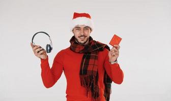 Headphones and red gift box. Young handsome man in New year clothes standing indoors against white background photo