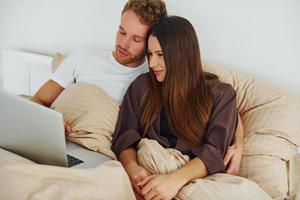 Couple lying down on bed with laptop. Interior and design of beautiful modern bedroom at daytime photo