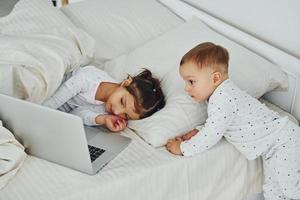 Little sister and brother uses laptop. Interior and design of beautiful modern bedroom at daytime photo