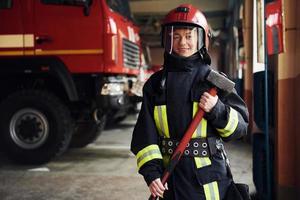 con martillo en las manos. bombero femenino en uniforme protector de pie cerca de camión foto