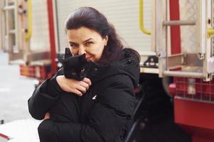 mujer feliz sostiene a su gato negro en las manos junto a un camión de bomberos al aire libre foto