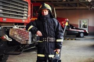 Holds axe in hands. Male firefighter in protective uniform standing near truck photo