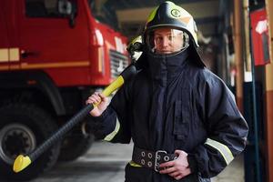 Holds axe in hands. Male firefighter in protective uniform standing near truck photo