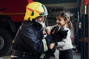 Cute black cat. Happy little girl is with male firefighter in protective uniform photo