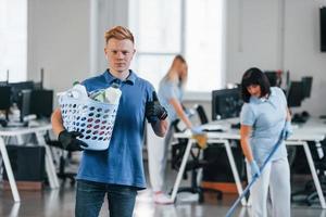 Man holds basket. Group of workers clean modern office together at daytime photo