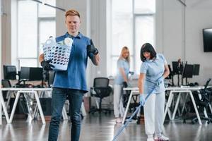 Man holds basket. Group of workers clean modern office together at daytime photo