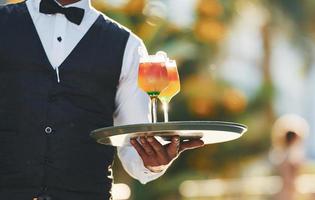 Holds cocktails. Black waiter in formal clothes is at his work outdoors at sunny daytime photo