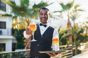 Holds cocktails. Black waiter in formal clothes is at his work outdoors at sunny daytime photo
