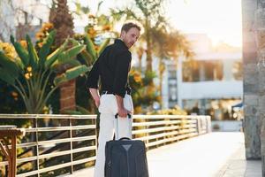 With travel bag. Young man is outdoors at sunny daytime. Concept of vacation photo