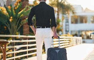 With travel bag. Young man is outdoors at sunny daytime. Concept of vacation photo