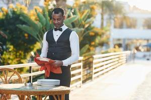 limpia los platos con un paño rojo. camarero negro con ropa formal está en su trabajo al aire libre durante el día soleado foto