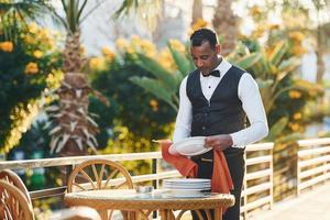 Wipes plates by red cloth. Black waiter in formal clothes is at his work outdoors at sunny daytime photo